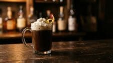 Buttsex cocktail in a clear glass coffee mug topped with whipped cream and a lemon twist on a dark wooden bar