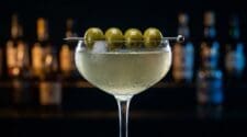 A Filthy Martini in a chilled coupe glass, cloudy pale olive-green liquid, four green olives on a pick across the rim, dark bar bokeh behind.