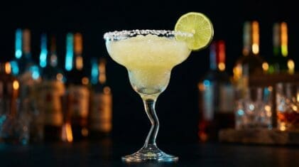 A Frozen Margarita in a classic margarita glass, pale yellow-green slushy texture, thick salt rim, fresh lime wheel garnish, dark bar bokeh with neon bottle accents.