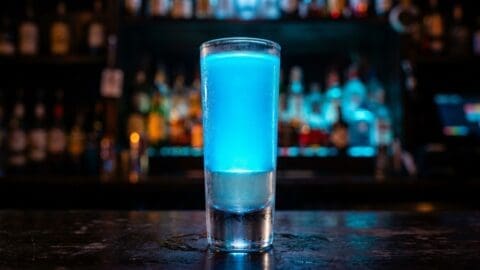 A Lightsaber Shot in a tall shot glass, bright electric blue liquid on top of a clear base, glowing against a dark bar background.