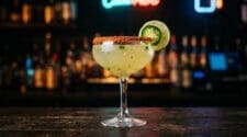 A Spicy Margarita in a classic coupe glass, cloudy pale yellow-green liquid, chili salt rim, fresh jalapeno slice and lime wheel garnish, dark moody bar background.