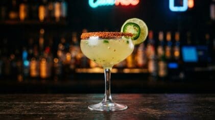A Spicy Margarita in a classic coupe glass, cloudy pale yellow-green liquid, chili salt rim, fresh jalapeno slice and lime wheel garnish, dark moody bar background.